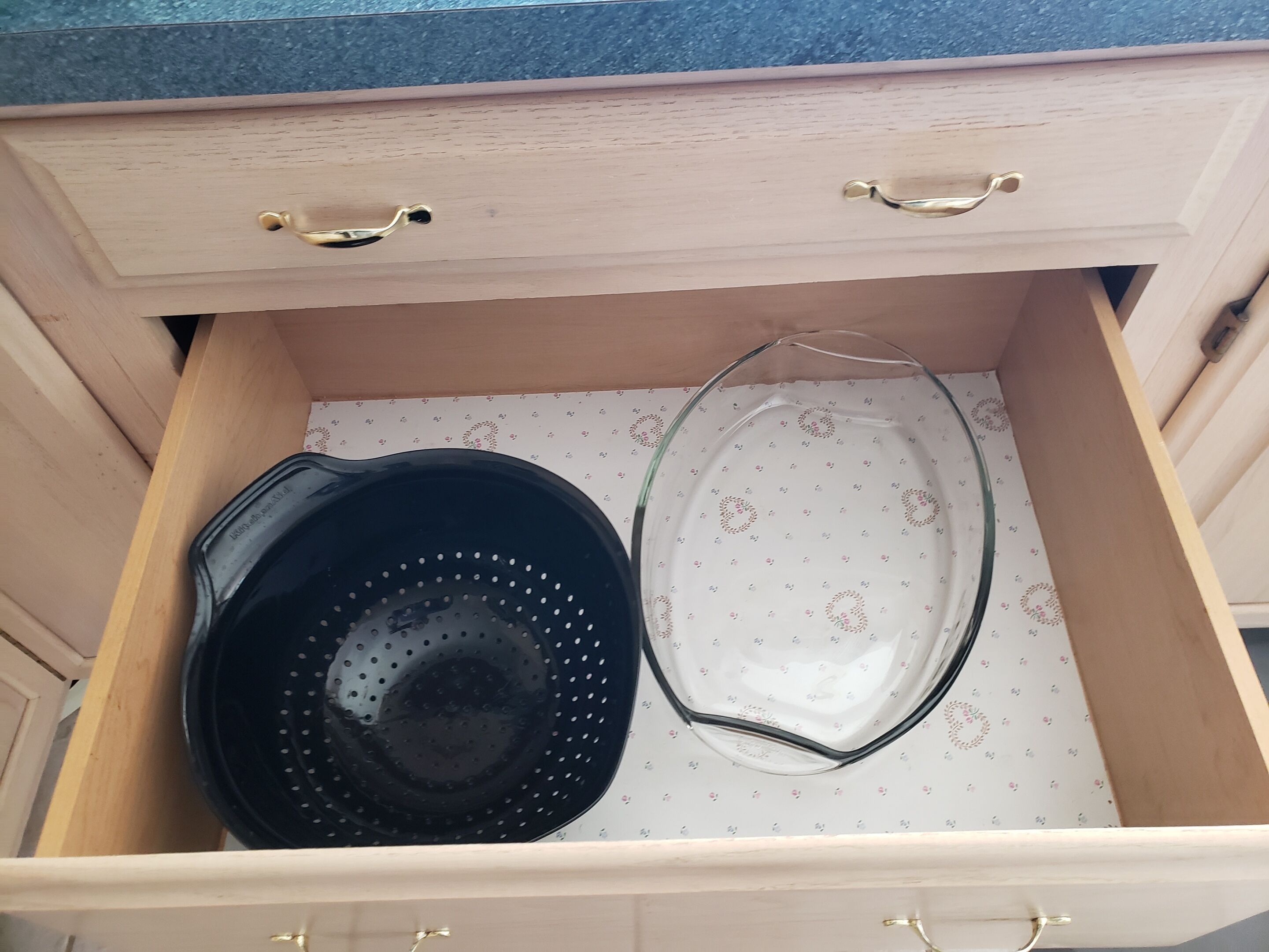 A drawer with two kitchen utensils: a black colander and a clear glass plate.