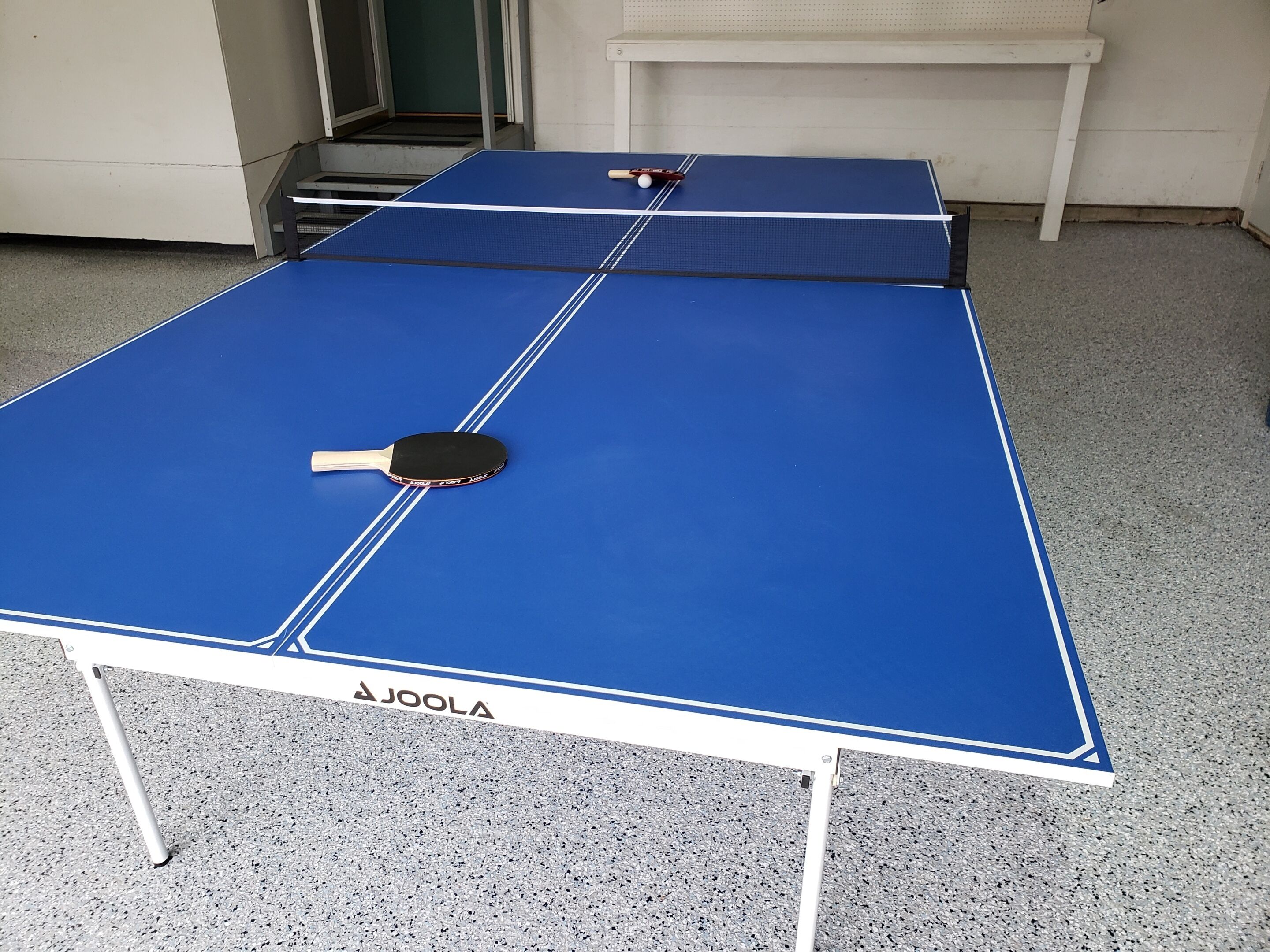 A blue ping pong table with a paddle, a white table leg, and a white bench in the background.