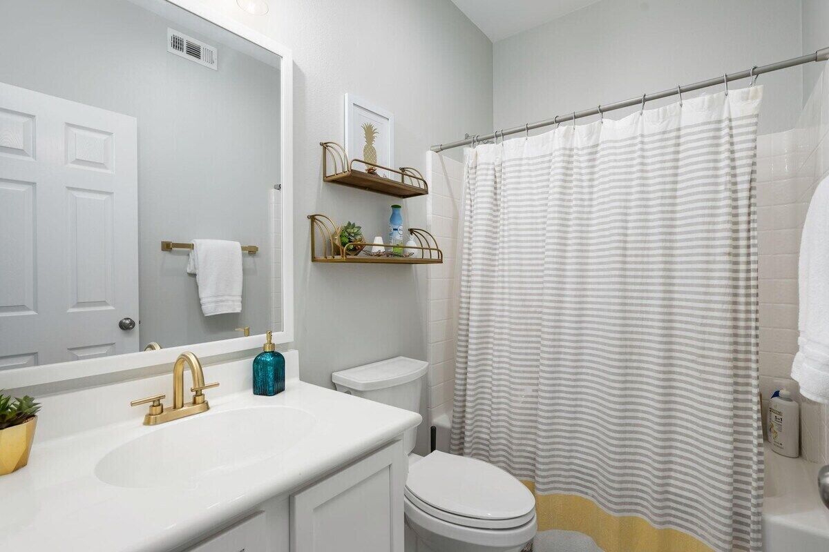 Un baño con lavamanos blanco, grifo dorado y un inodoro. Hay repisas sobre el inodoro y una cortina de ducha con rayas horizontales.