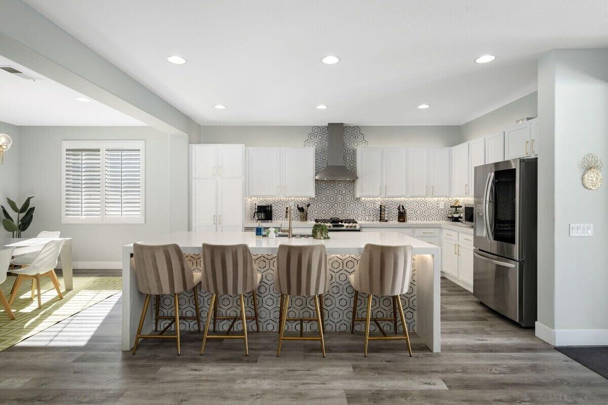 A modern kitchen with white cabinetry, a central island with bar stools, a stainless steel refrigerator, and a patterned backsplash.