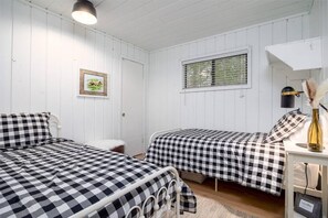 Ein Zimmer mit zwei Betten, weißen Wänden, einem Fenster mit Jalousien, einem gerahmten Bild und einer Lampe auf einem Nachttisch.