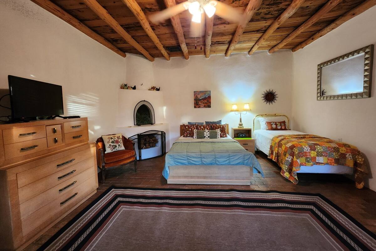 Un dormitorio con techo de madera, una cama con colcha colorida, un televisor sobre un tocador de madera y una chimenea.