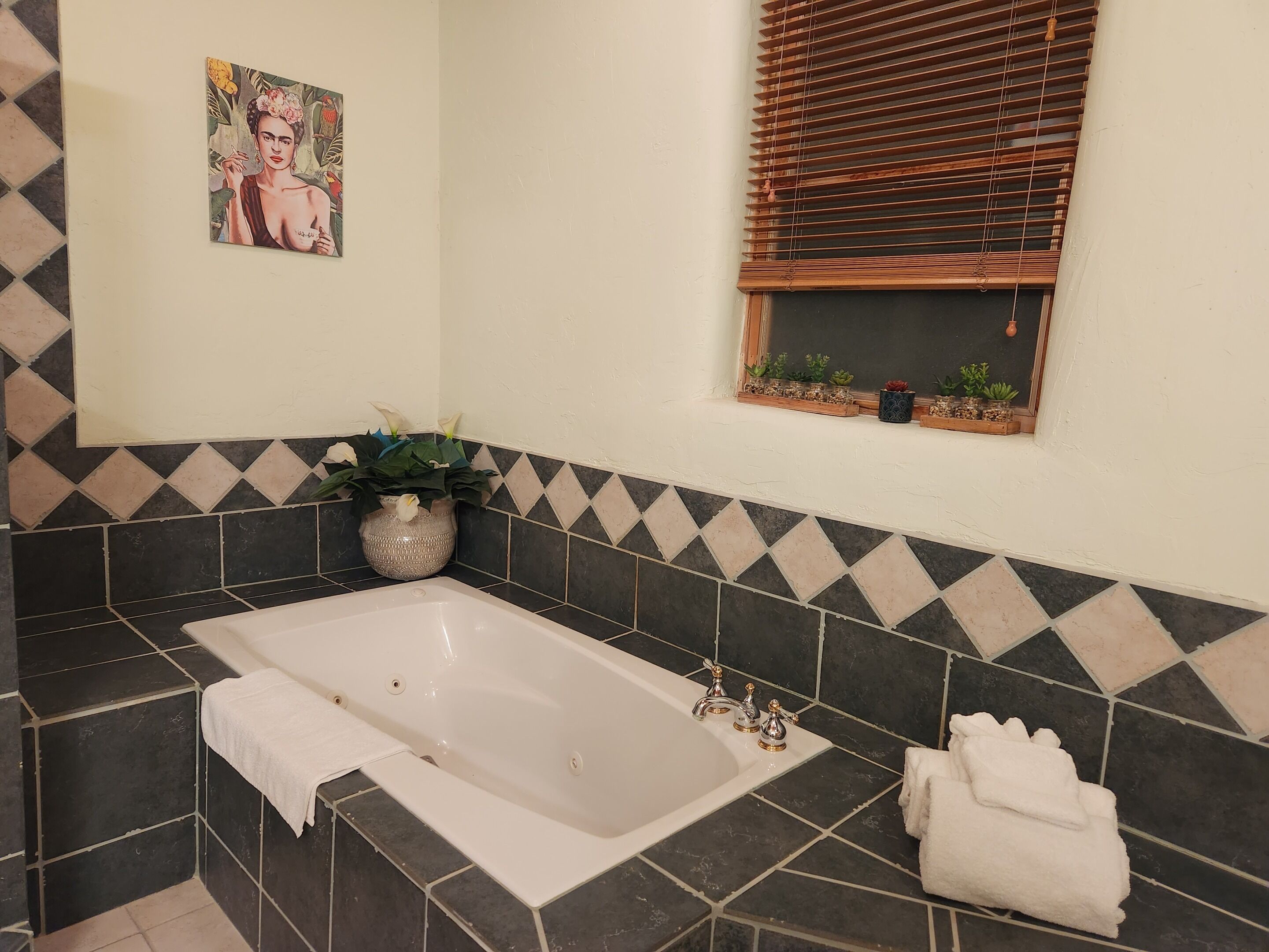 Ein Badezimmer mit Badewanne, einem Blumentopf, einem Fenster mit Jalousien und einer dekorativen Fliesenwand.