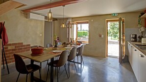 Un comedor con mesa de madera, sillas e iluminación colgante. Se aprecia una pared de ladrillo y una puerta que da al exterior.