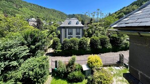 Un grand bâtiment en pierre, avec un toit gris, entouré d’une végétation luxuriante et avec des collines en arrière-plan.