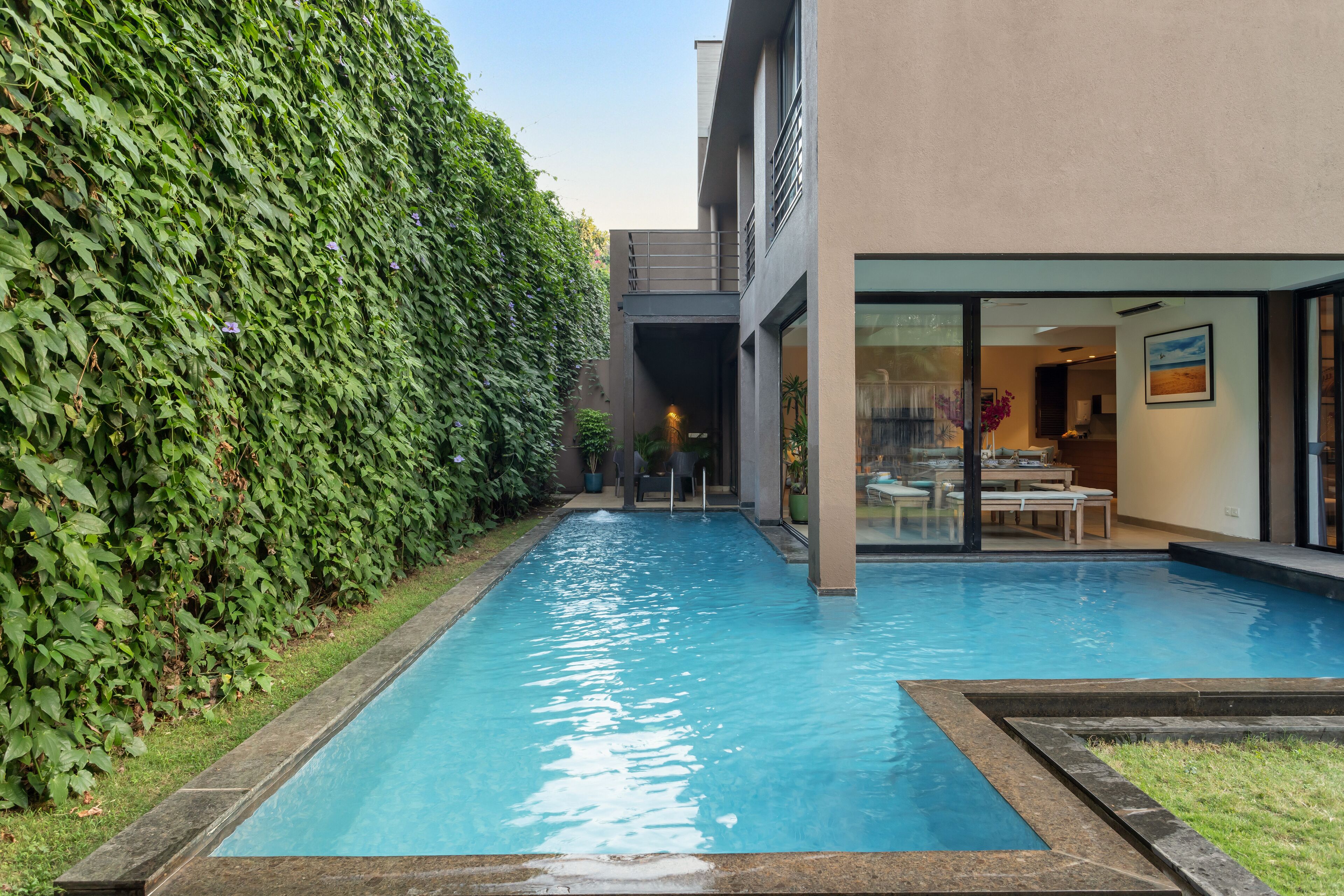 A swimming pool with a stone border, surrounded by a lush green hedge, and a modern building with a glass door entrance.