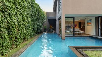 A swimming pool with a stone border, surrounded by a lush green hedge, and a modern building with a glass door entrance.