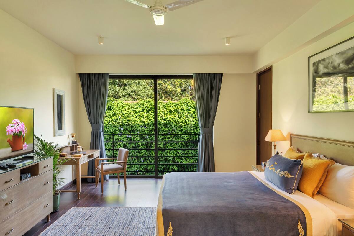 A hotel room with a bed, desk, chair, TV, and a view of greenery through the window.