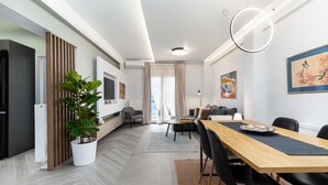 A modern living room with a dining area, featuring a wooden dining table, black chairs, a potted plant, and a large painting on the wall. - Amazonia Apt by halu! (Thessaloniki)
