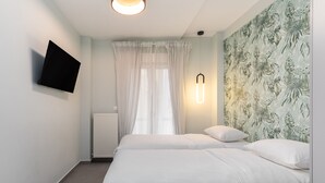 A bedroom with a bed, a TV mounted on the wall, a window with sheer curtains, and a wall with a tropical leaf pattern. - Amazonia Apt by halu! (Thessaloniki)