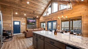 A spacious kitchen with granite countertops, wooden cabinets, and a large island. There is a dining area with a brown leather couch and a fireplace. The room has wooden walls and large windows.