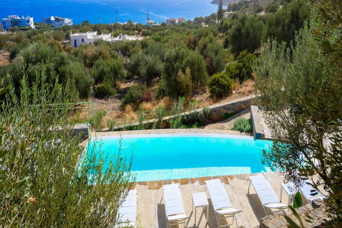 A pool with lounge chairs, surrounded by greenery and overlooking a coastal landscape with buildings.