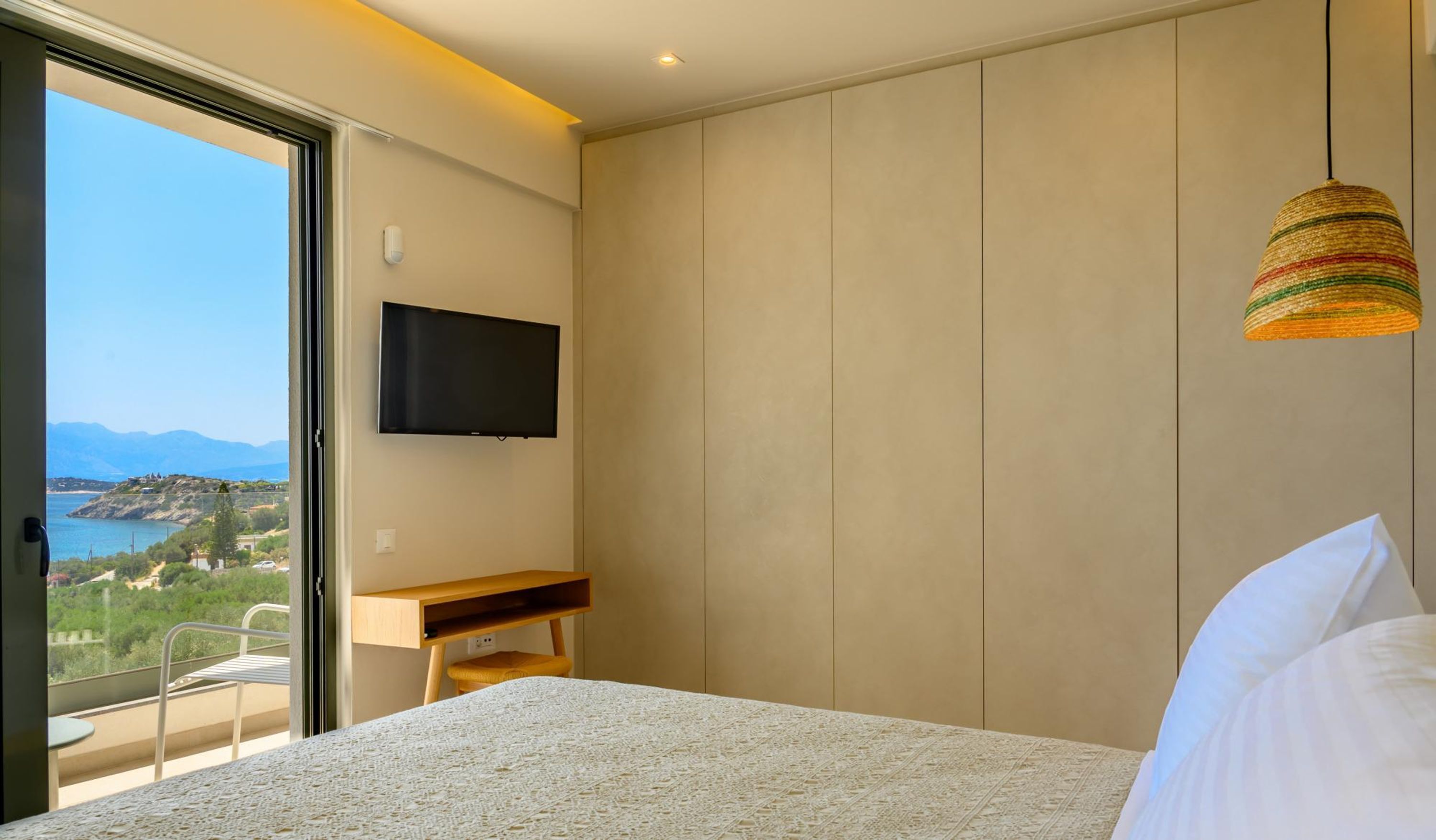 A bedroom with a bed, a desk, a TV, and a balcony with a view of the sea and mountains.