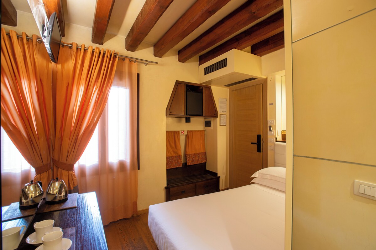 A hotel room with a bed, a wooden table with cups and a kettle, a TV, and a wardrobe.
