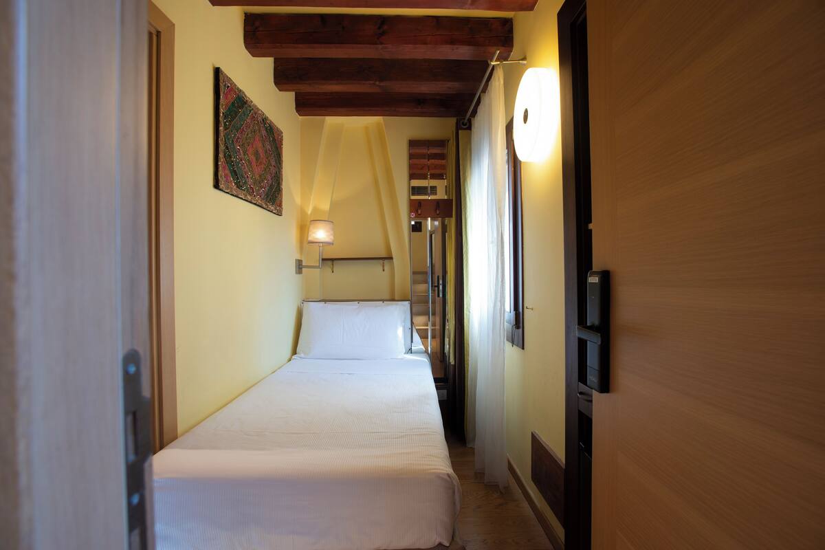 A narrow hallway with a bed at the end, wooden ceiling beams, a wall-mounted lamp, and a door on the left.
