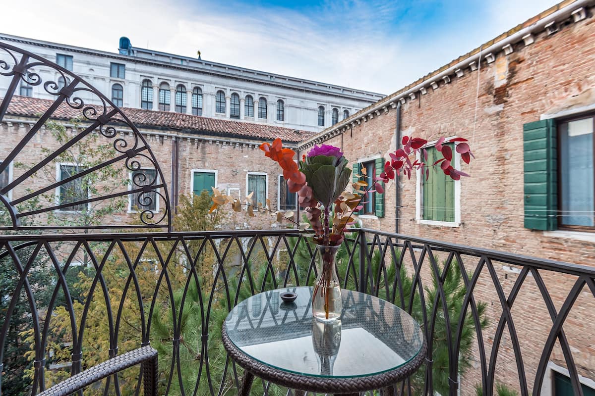 A balcony with a glass table and a vase of flowers, overlooking a historic building with brick walls and green shutters.