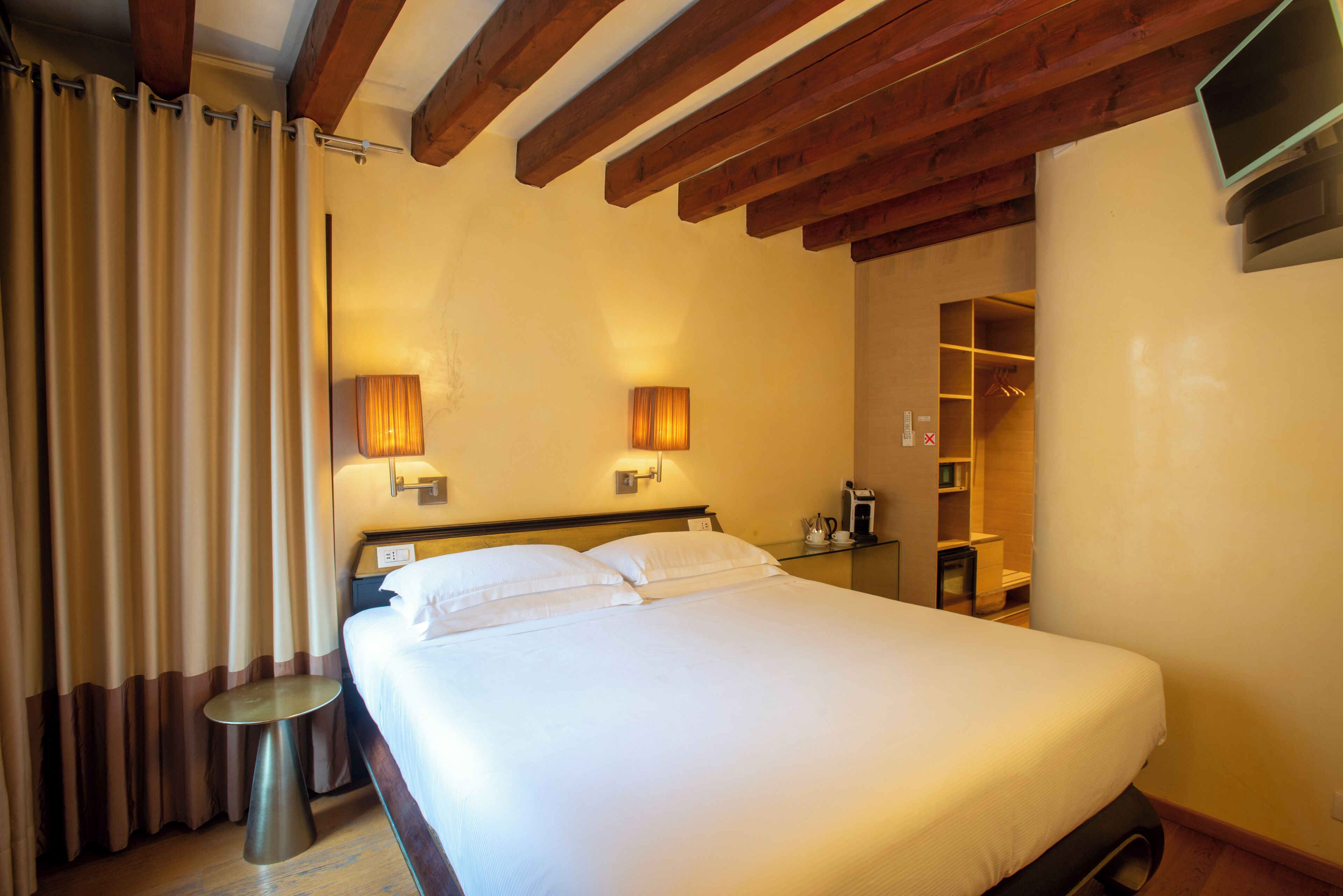 A hotel room with a large bed, wooden ceiling beams, a bedside table, and a wardrobe.