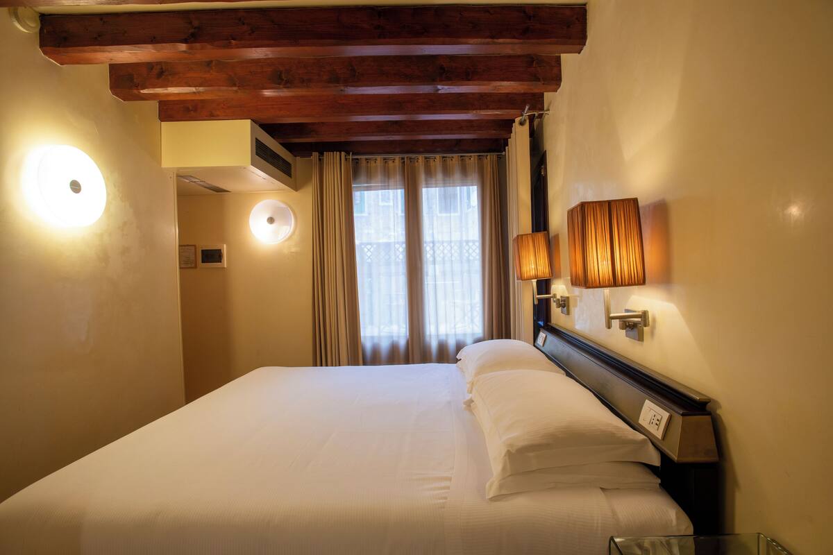 A hotel room with a wooden ceiling, a bed with white linens, a nightstand with a lamp, and a window with curtains.