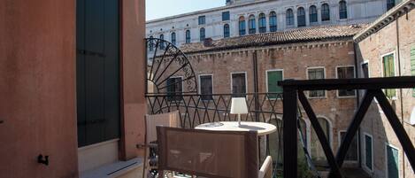 A balcony with a table and chair, a window with a green shutter, and a building with arched windows in the background.
