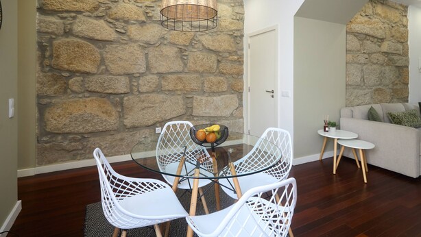 A dining area with a glass table and white chairs, a stone wall, and a sofa in the background. - Oporto Yellow Villas 7 (Porto)