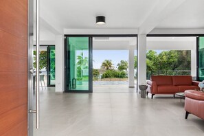 A spacious modern living area with a large sliding glass door leading to a balcony, a red leather sofa, and a minimalist coffee table. - Salty Villa | Modern Beachfront Oasis (Arecibo)