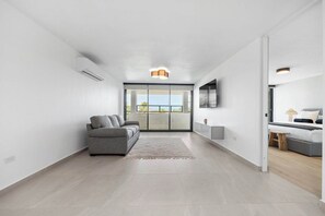 A modern living room with a grey sofa, a flat-screen TV mounted on the wall, and a large window providing a view of the outdoors.