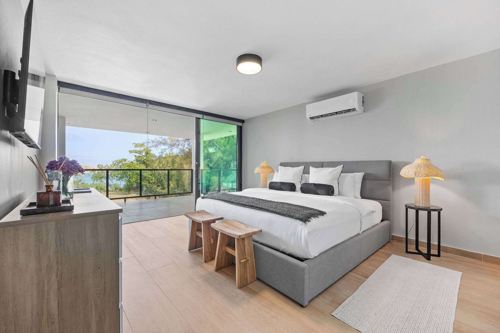 Ein modernes Schlafzimmer mit einem großen Bett, Nachttischlampen, einem kleinen Holzschemel und Blick durch eine Glastür.