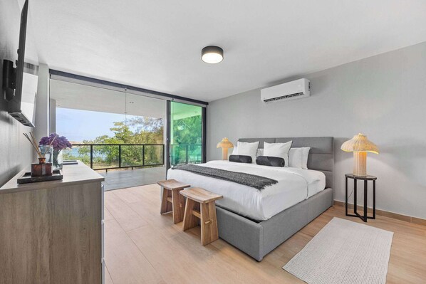 A modern bedroom with a large bed, bedside lamps, a small wooden stool, and a view through a glass door.