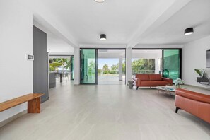 A spacious living area with a sofa, a bench, and a coffee table. There is a sliding glass door leading to an outdoor area with a view of greenery and a pool. - Salty Villa | Modern Beachfront Oasis (Arecibo)