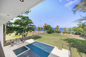 A swimming pool with a view of the sea, a grassy lawn, and a wooden deck with outdoor furniture.