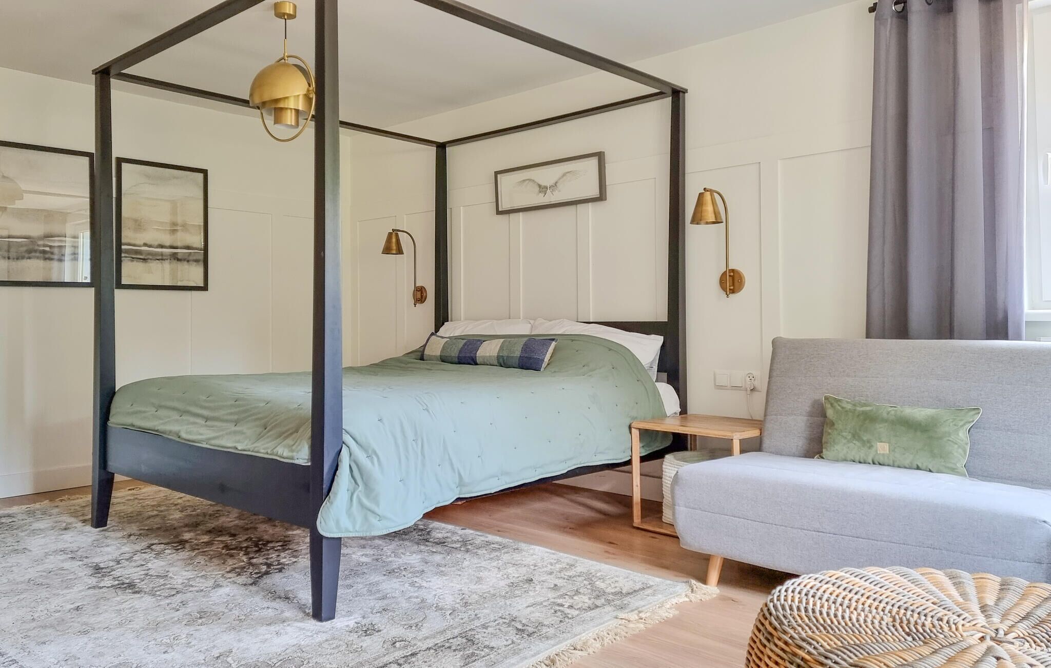 A four-poster bed with a green bedspread and pillows, a grey sofa, a small wooden nightstand, and a woven ottoman.