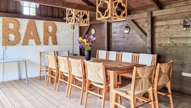 Un comedor rústico de madera con una mesa larga y sillas de mimbre, un cartel grande que dice 'BAR' y un jarrón con flores.