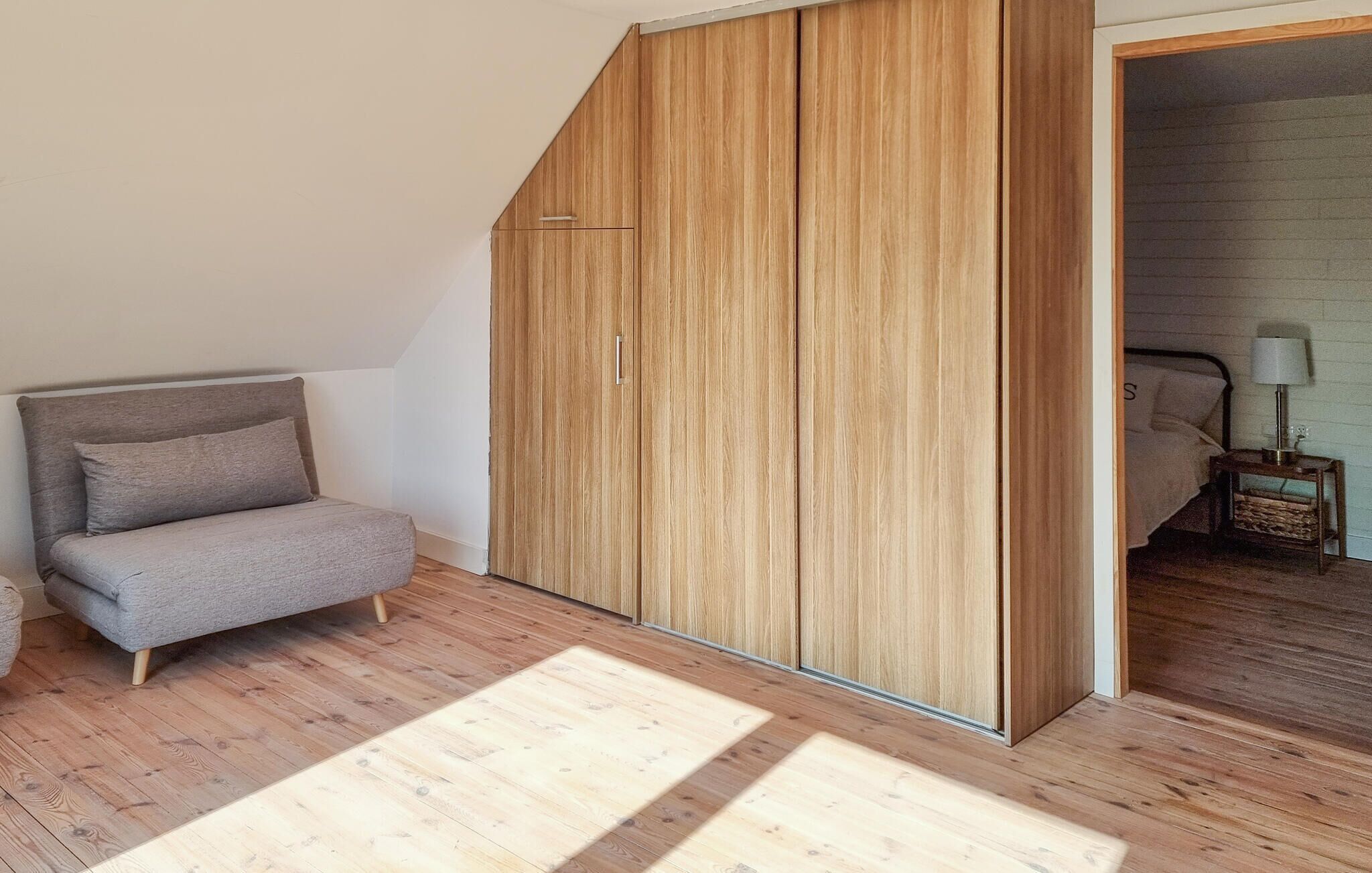 A modern bedroom with a grey sofa, wooden wardrobe, and a small bedside table with a lamp.
