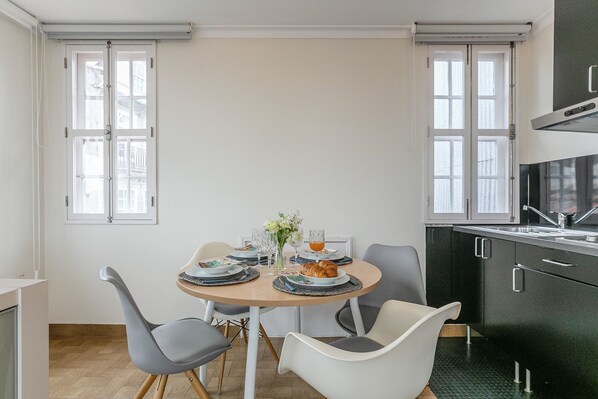 Un coin repas avec une table ronde, deux chaises, une cuisine avec des meubles noirs et une fenêtre donnant sur des immeubles.