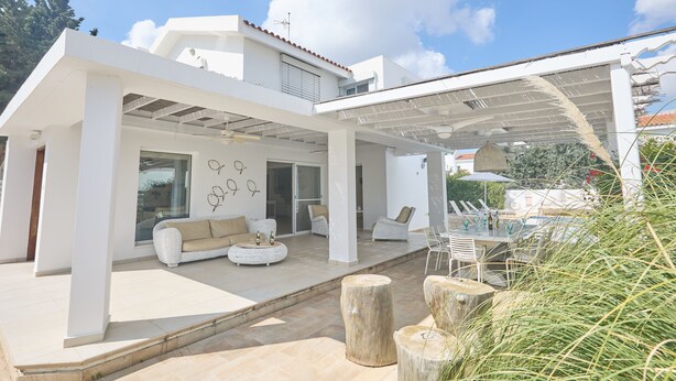 A modern outdoor living space with a white exterior, a covered patio area, and a dining setup. - Protaras Serifos Villa Sv07 (Paralimni)