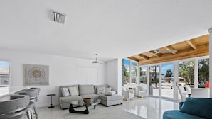 Una sala de estar moderna con un sofá gris, un sillón verde mar y una mesa de centro de vidrio. Una ventana grande ofrece vistas a la playa.