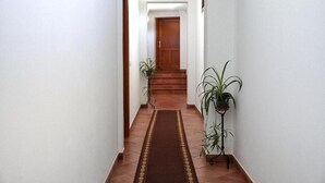 Un couloir se termine par une porte en bois, de part et d’autre se trouvent des plantes en pot, et un tapis à motifs recouvre le sol.