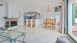Un vaste espace de vie comprenant une table basse en verre, une cheminée, une cuisine avec des meubles blancs et un coin repas avec des chaises en bois.
