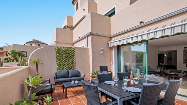 Une terrasse extérieure avec une table à manger, des chaises et un canapé. Un mur est recouvert d’une plante grimpante, et une petite plante en pot y est également placée.