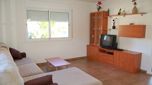 Un salon avec un canapé, une petite table, un téléviseur posé sur un meuble en bois et une fenêtre avec des stores.