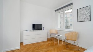 A modern, minimalistic room with a herringbone wooden floor, a flat-screen TV on a white dresser, two chairs, and a large window with a view of buildings. - Stylish Studio near the Fair | Private Parking (Poznan)