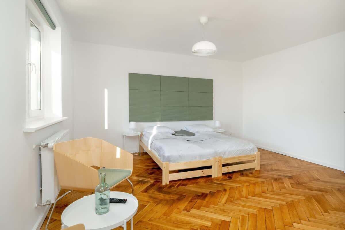 A bright bedroom with a double bed, a green upholstered headboard, and a small bedside table with a water bottle.
