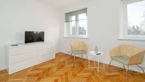 A modern, minimalistic room with a herringbone wood floor, white walls, a flat-screen TV on a white cabinet, two chairs with green cushions, and a small round table with a bottle. - Functional Studio in Poznan | Parking, SmartTV (Poznan)