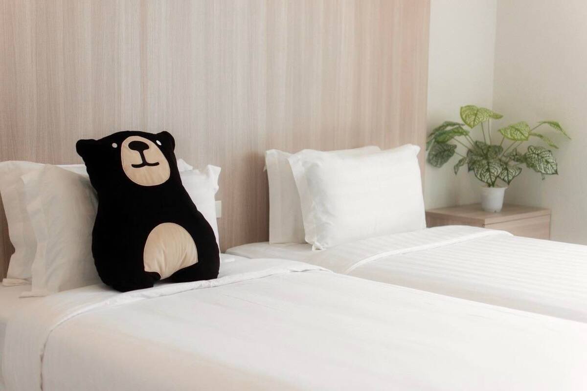 A hotel room with a bed, white bedding, a black bear-shaped pillow, a white pillow, a wooden headboard, a bedside table with a plant, and a white nightstand.