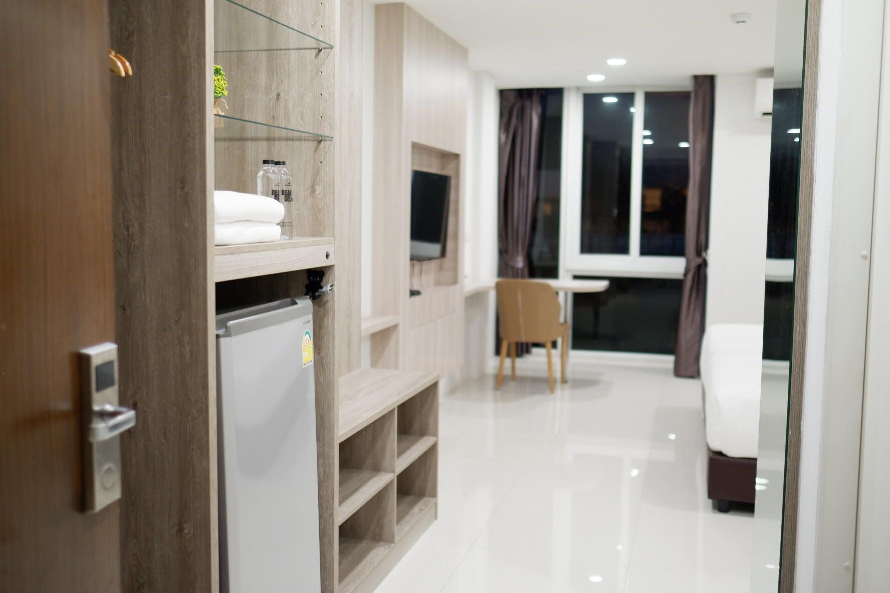 A modern hotel room with a wooden wardrobe, a small refrigerator, a TV mounted on the wall, a dining table with a chair, and a large window with curtains.