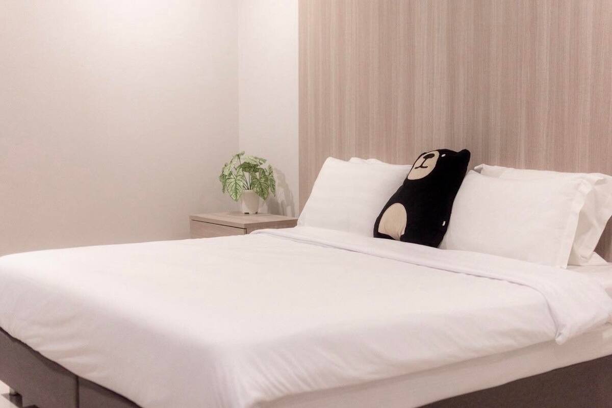 A neatly made bed with white linens, a small bedside table with a plant, and a wall with vertical blinds.