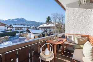 Un balcon avec une table en bois, des chaises et un canapé. On y aperçoit un paysage enneigé et des bâtiments en arrière-plan.