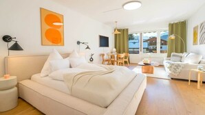 Een moderne slaapkamer met een groot bed, nachtkastjes, een bureau en een stoel. Er is een raam met gordijnen, een plafondlamp en kunst aan de muur.