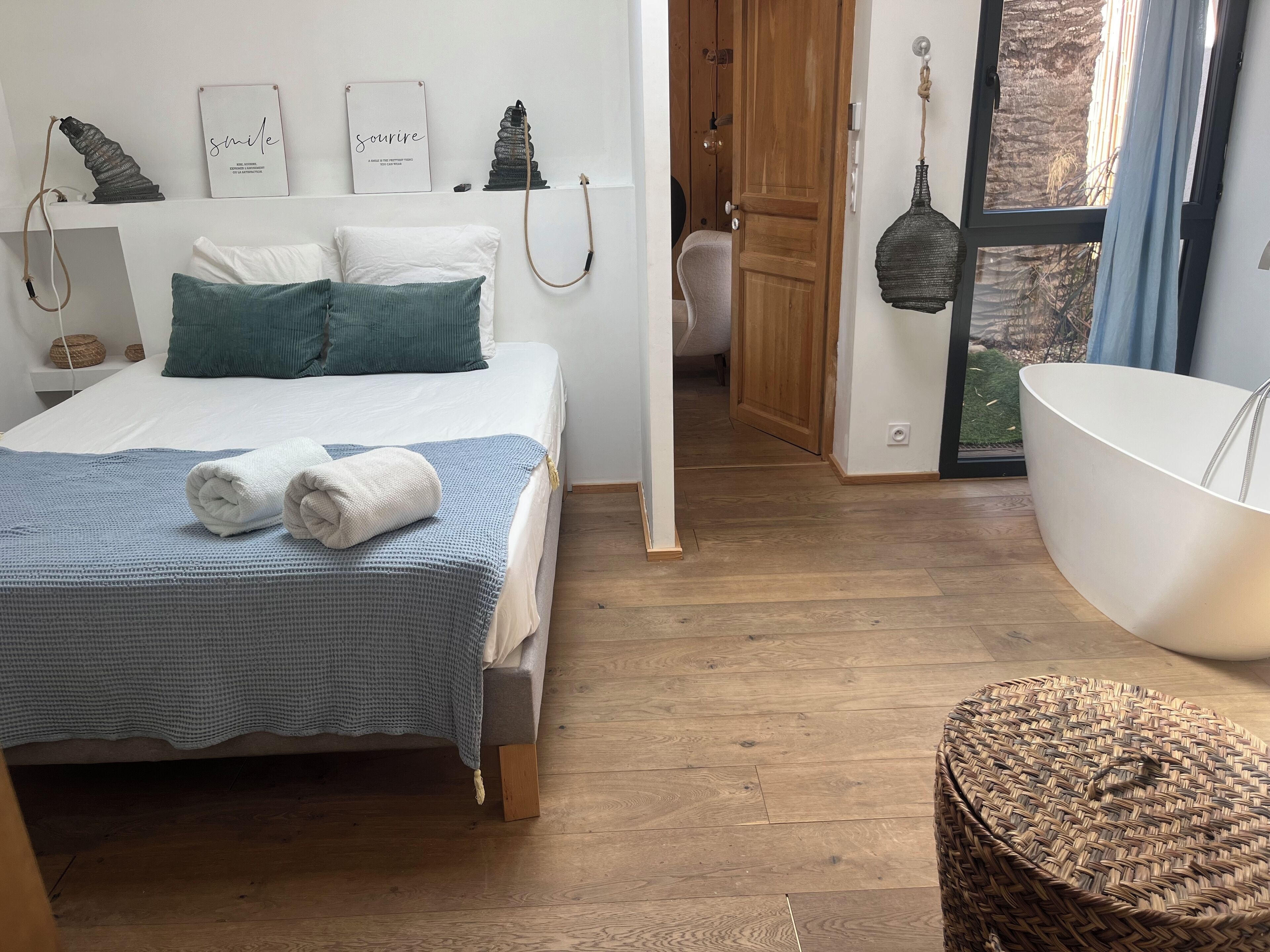Un dormitorio con una cama, un ventanal grande, piso de madera, una bañera blanca y un taburete tejido.