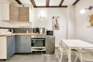 Une cuisine compacte avec des meubles gris, un four encastré, une machine à café et un four à micro-ondes. On y trouve également un petit coin repas avec une table et des chaises blanches.